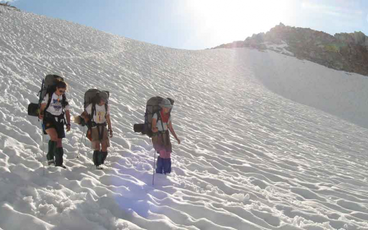 three people wearing backpacks hike down a snowy slope on an outward bound course